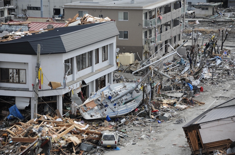 2. A Fishing Boat Sits Ashore Next to A Building In Ofunato, March 15, 2011, Nippon-koku (Nihon-koku) - Japan. Photo Credit: Mass Communication Specialist 1st Class Matthew M. Bradley, United States Navy; Defense Visual Information (DVI, http://www.DefenseImagery.mil, 110315-N-KK192-118) and United States Navy (USN, http://www.navy.mil, 110315-N-2653B-118, http://www.navy.mil/view_image.asp?id=98448, https://www.navy.mil/management/photodb/photos/110315-N-2653B-118.jpg), United States Department of Defense (DoD, http://www.DefenseLink.mil or http://www.dod.gov), Government of the United States of America (USA).