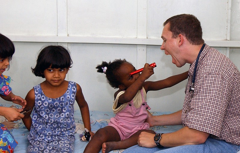 With a Flashlight In Her Right Hand, a Little Girl From St. Ann's Girls Orphanage Checks the United States Air Force (USAF) Captain's Mouth, July 2004, Georgetown, Co-operative Republic of Guyana. Photo Credit: SSgt. Srayuth Pinthong, 1st Combat Camera Squadron, 820th RED HORSE Squadron, United States Air Force; Defense Visual Information Center (DVIC, http://www.DoDMedia.osd.mil, DFSD0700859 and 040703F6335P006) and United States Air Force (USAF, http://www.af.mil), United States Department of Defense (DoD, http://www.DefenseLink.mil or http://www.dod.gov), Government of the United States of America (USA).